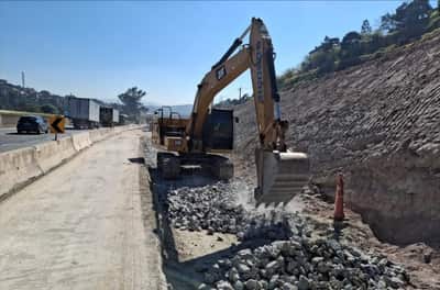 Escavadeira hidráulica movimentando enrocamento na faixa adicional em implantação, junto à barreira de concreto que separa o tráfego
