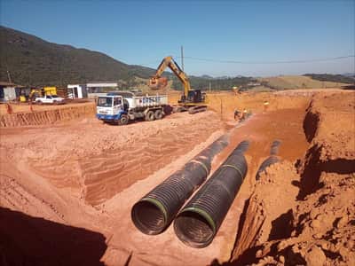 Tubulação corrugada de drenagem posicionada em vala, com escavadeira e caminhão basculante ao fundo