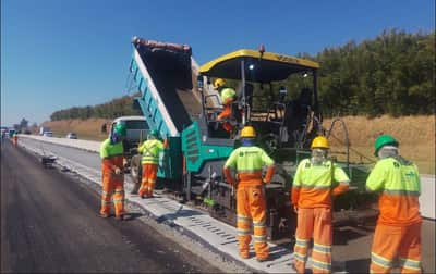 Caminhão basculante descarregando massa asfáltica na vibroacabadora, acompanhado pela equipe de pavimentação