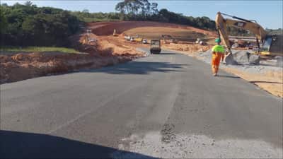 Camada asfáltica recém-aplicada na perimetral, com trabalhador uniformizado e escavadeira na lateral da pista