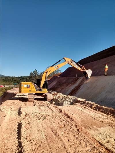 Escavadeira hidráulica carregando material no corte do terreno, com o talude escalonado ao fundo